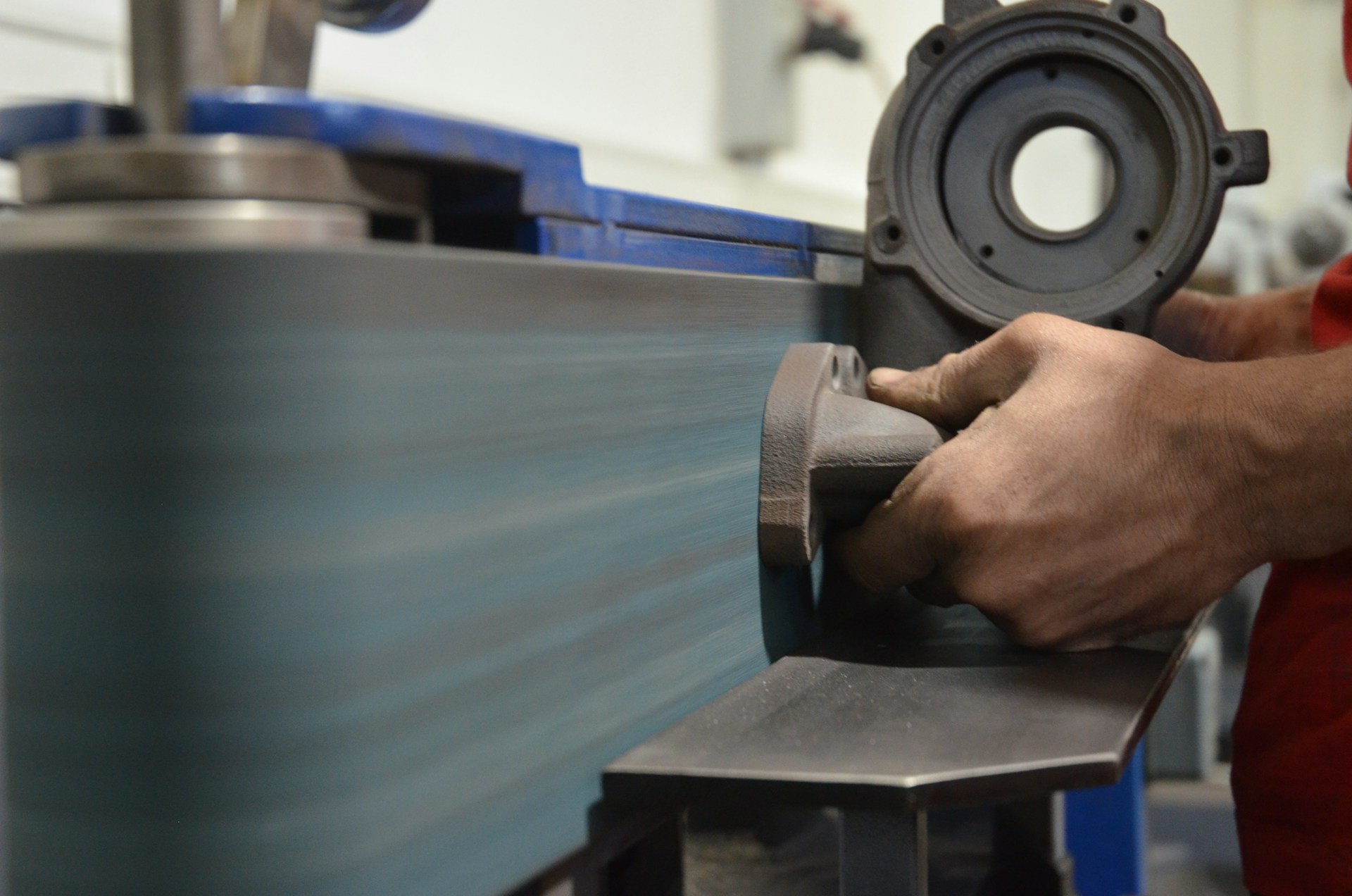 A person is holding a metal part to a sander for surface preparation before coating